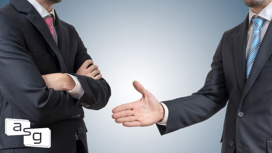 Two businessmen engaging, one with arms crossed, the other extending a handshake, representing the challenge of an uncoachable employee resistant to teamwork and feedback as described in a blog on the importance of coachability in the workplace