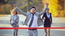 A jubilant man dressed in a business suit with a tie is breaking through a red finish line tape, arms raised in victory as the top seller, with a focused group of businesswomen running behind him. This image humorously represents Dave Reynolds' achievement as the Top Seller for the third year in a row, as described in the satirical blog post set in the competitive sales environment