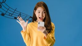Excited young woman in a yellow sweater looking at her smartphone with musical notes flowing from the screen, symbolizing the joy of receiving a personalized song as a novel sales engagement tactic, as described in a satirical blog post about unique client outreach methods.