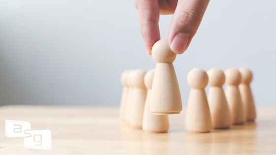 A person's hand selecting a single game piece from a line of identical pieces, symbolizing the importance of proactive leadership in sales management. This image illustrates the concept of choosing the right strategy and personnel, much like a sales manager must select the best course of action and team members to avoid reactive management and ensure success.