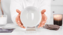 A person in a white shirt holds their hands around a crystal ball on a table, symbolizing the uncertainty and guesswork often associated with sales forecasting. Nearby are candles, cards, and cloth, reinforcing the theme of prediction and unclear outcomes. The image reflects the unpredictability of sales pipelines and the need for structured forecasting models.