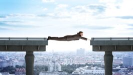 A businessman in a suit stretches his body horizontally across a gap between two unfinished bridge sections, acting as a human connection. Below him, a cityscape with buildings and streets extends into the distance.