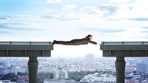 A businessman in a suit stretches his body horizontally across a gap between two unfinished bridge sections, acting as a human connection. Below him, a cityscape with buildings and streets extends into the distance.