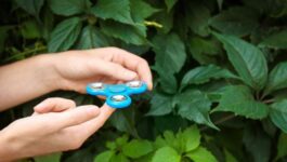 A person’s hands holding a blue fidget spinner against a background of lush green leaves. The fingers are positioned as if about to spin the toy, which has three rounded edges with metallic bearings in the center of each. The vibrant greenery contrasts with the bright blue of the fidget spinner.