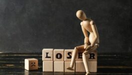 A wooden artist mannequin sits hunched over on a row of wooden blocks that spell out "LOSER," with its head tilted downward in a dejected posture. To the left, an additional wooden block features a sad face emoji. The dark background and moody lighting emphasize a theme of failure or discouragement.