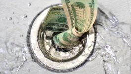 A rolled-up stack of U.S. twenty dollar bills being sucked down a stainless steel sink drain with water swirling around.