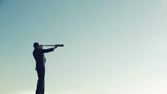 A business professional in a suit stands on a ledge using a handheld telescope to look into the distance. The clear sky background emphasizes focus and visibility. The image represents sales forecasting and forward-looking revenue projections based on verified buyer input, qualification standards, and opportunity scoring—key elements of the Forecast Layer in the Revenue S.P.E.E.D.™ Model.