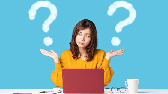 Woman in yellow sweater sitting at a desk with a red laptop, looking confused with both hands raised, surrounded by cloud-shaped question marks on a blue background—representing uncertainty or indecision.