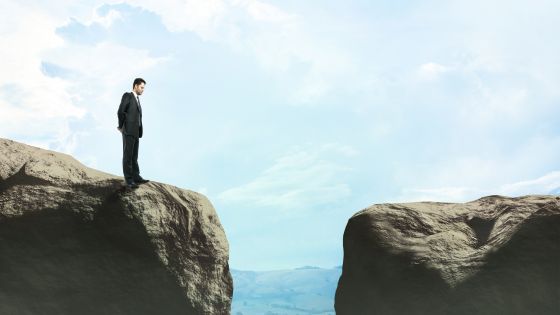 A businessperson in a suit stands on the edge of a cliff, staring across a wide gap toward another rocky ledge, symbolizing the uncertainty and risk of unreliable sales forecasts.